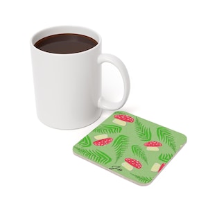 May include: A white ceramic mug filled with dark liquid sits beside a square coaster. The coaster has a light green background with a pattern of red mushrooms and green fern leaves. The mug has a large handle.