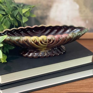 May include: A vintage ceramic bowl with a scalloped edge and a decorative floral design in shades of green, pink, and brown. The bowl is sitting on a stack of black books.