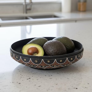 May include: A round black bowl featuring a decorative pattern of small white, gold, and brown dots arranged in scalloped arches along the exterior rim. The bowl contains two whole dark green avocados and one halved avocado with a visible brown pit.