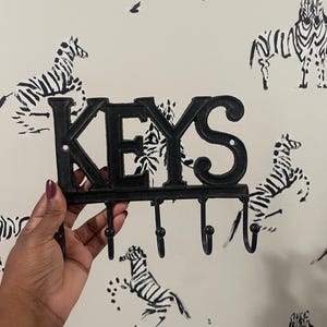 May include: Black metal key holder with the word "KEYS" in large, stylized letters. The key holder has five hooks below the letters. The background is a light-colored wall with a repeating zebra pattern.