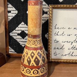 May include: A tall, decorative vase with a cylindrical neck and a bulbous base. The vase features a beige background with red and brown geometric patterns. The top and bottom of the vase are red. The vase is displayed on a wooden surface.