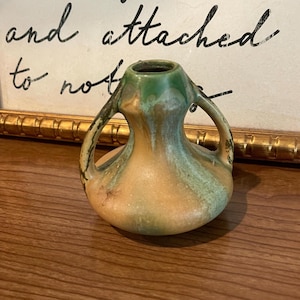 May include: A small ceramic vase with two handles, featuring a rounded base and a narrow neck. The surface has a blended glaze finish in shades of muted green, tan, and brown, with vertical drips of color running down the sides of the vessel.