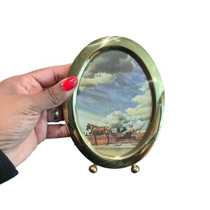 May include: Oval brass picture frame with a landscape scene featuring a horse-drawn carriage under a cloudy sky. The frame has a polished finish and small ball feet.