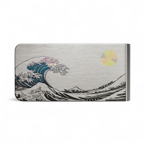 以下が含まれることがあります： 日本の波のデザインとカラフルな太陽が特徴のシルバーマネークリップ。アートワークには、白い波頭を持つ大きな青い波と小さなボートが含まれています。クリップは角が丸い長方形です。