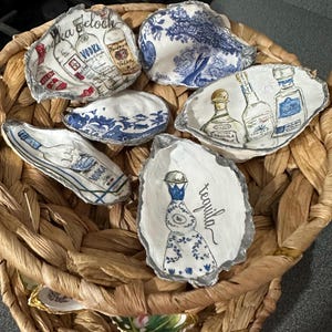 May include: A collection of painted oyster shells, each featuring a different liquor bottle design. The shells are decorated with images of vodka and tequila bottles, with text labels. The shells are arranged in a woven basket.