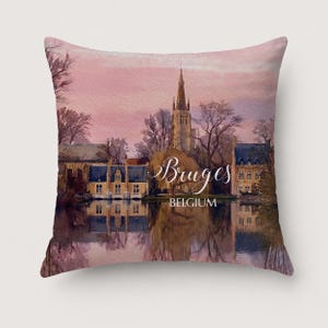 May include: A square throw pillow featuring a watercolour-style illustration of a cityscape in Bruges, Belgium. The scene depicts historic buildings and a church spire reflected in water under a soft pink sky, with the text Bruges BELGIUM printed.