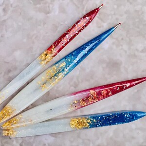 Puede incluir: Cuatro bolígrafos hechos a mano con purpurina y detalles de pan de oro. Los bolígrafos tienen una base blanca con puntas de purpurina roja y azul. Las puntas son metálicas y los bolígrafos están decorados con pan de oro.