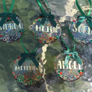 Puede incluir: Cinco adornos navideños redondos y transparentes con nombres en escritura blanca: Andrea, Heidi, Sofia, Mitchell y Angel. Cada adorno tiene purpurina colorida, una cinta verde y un lazo verde. Los adornos cuelgan sobre una superficie reflectante.