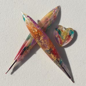 May include: Two colorful, handmade knitting needles crossed with a small heart-shaped charm. The needles and charm are filled with vibrant hues of red, orange, yellow, purple, and green, accented with gold flakes. The needles are approximately 7 inches long.
