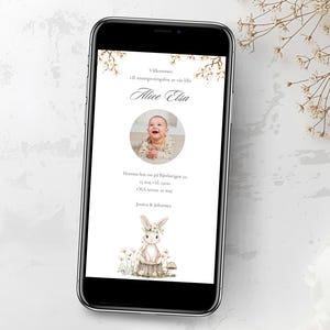 May include: A smartphone displays a baby announcement with the name "Alice Elsa" and a photo of a smiling baby. The design includes floral accents and a cartoon bunny illustration. The phone rests on a white surface.