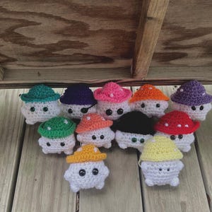 Puede incluir: Una colección de once figuras de setas de ganchillo con cuerpos blancos y gorros coloridos. Los gorros son de varios colores, incluyendo verde azulado, morado, rosa, naranja, negro, rojo y amarillo. Cada seta tiene dos ojos negros de botón.