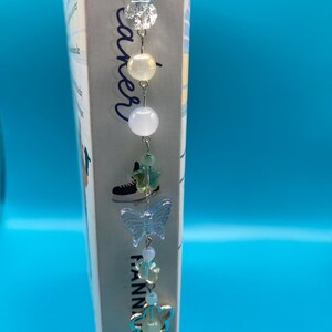 May include: A handmade beaded bookmark with a clear butterfly charm and various beads in white, green, and iridescent colors. The bookmark is attached to the spine of a book with the title "Breaker" and the author's name "Hanna Grace".