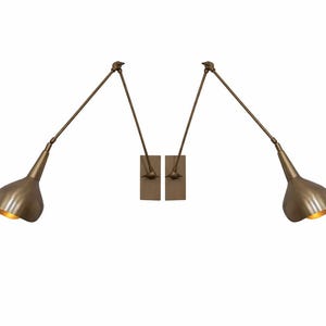 May include: Two antique brass wall sconces with adjustable arms and cone-shaped shades. The sconces are mounted on rectangular backplates and have a warm, inviting glow. The design is reminiscent of mid-century modern lighting.
