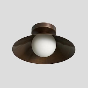 May include: A bronze-colored ceiling light fixture with a round, white globe bulb. The fixture has a circular base and a wide, downward-facing shade. The light is designed to be mounted on a ceiling.