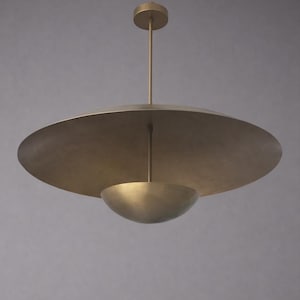 May include: A modern brass pendant light fixture. The light features a large, flat, circular shade above a smaller, bowl-shaped element. A slender rod extends from the ceiling to support the fixture, creating a minimalist design.