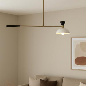 May include: A modern, brass-toned ceiling light fixture with a black and white shade. The light has a horizontal arm with a black handle on one end and a white, dome-shaped shade with a lightbulb on the other. The fixture is suspended above a beige sofa.