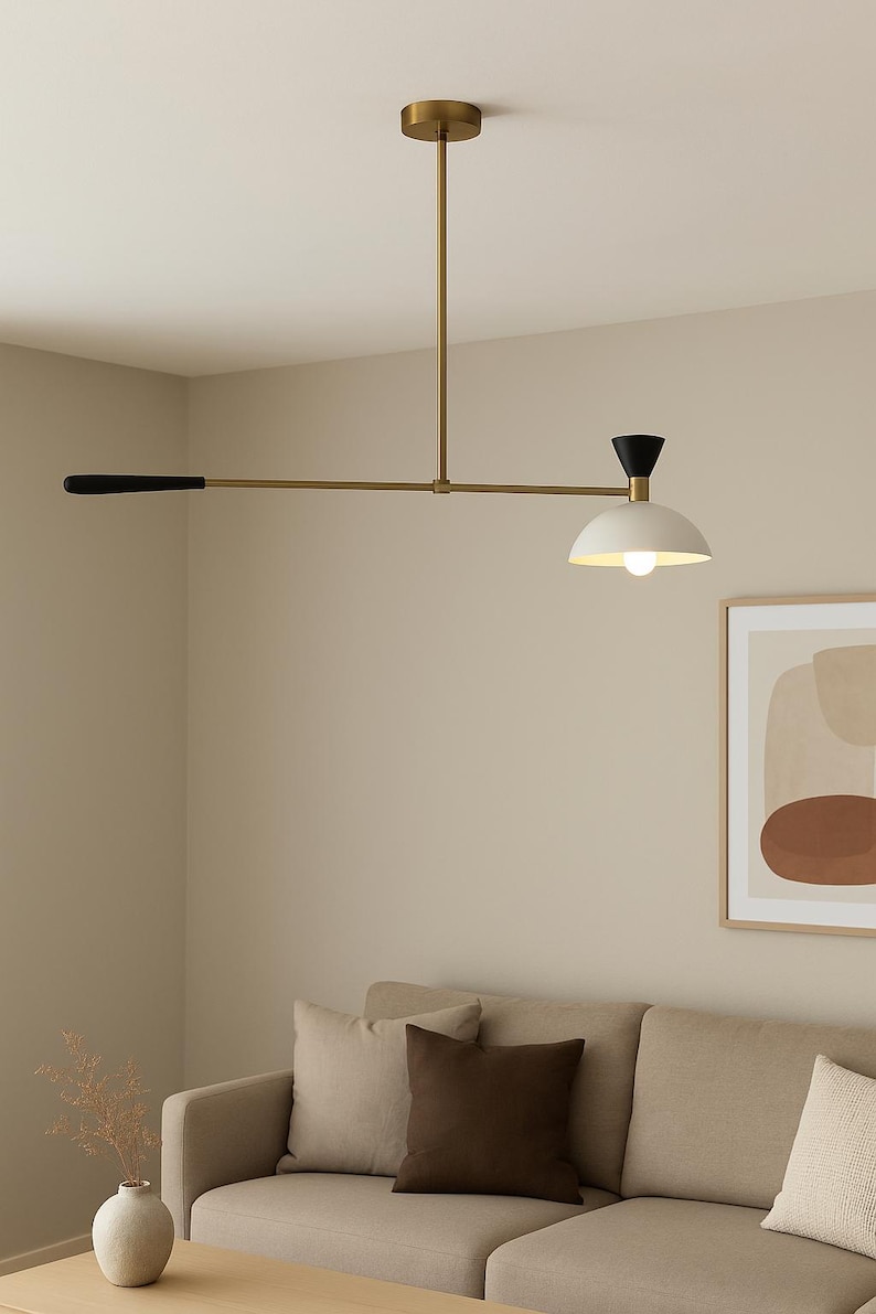 May include: A modern, brass-toned ceiling light fixture with a black and white shade. The light has a horizontal arm with a black handle on one end and a white, dome-shaped shade with a lightbulb on the other. The fixture is suspended above a beige sofa.