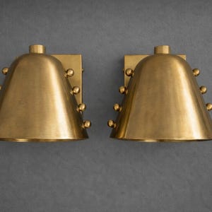 May include: Two matching brass wall sconces. Each sconce has a bell-shaped shade with a series of small, round decorative accents. The sconces are mounted on rectangular backplates, and the finish is a warm, metallic gold.