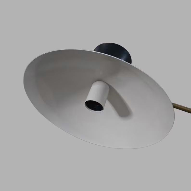 May include: A modern, minimalist lamp with a white, oval-shaped shade. The lamp features a black base, a white cylindrical light fixture, and a brass-colored arm. The design is sleek and contemporary, suitable for various interior styles.