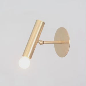 May include: A brass-colored wall-mounted light fixture. The fixture features a cylindrical light head, an adjustable arm, and a circular base. The light bulb is visible at the end of the cylinder. The design is minimalist and modern.