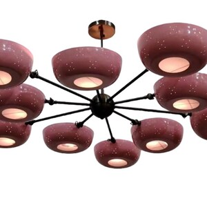 May include: A modern chandelier with ten dusty rose-colored, bowl-shaped shades. Each shade has a speckled pattern and emits a warm light. The fixture has a black frame and a brass-colored ceiling mount.