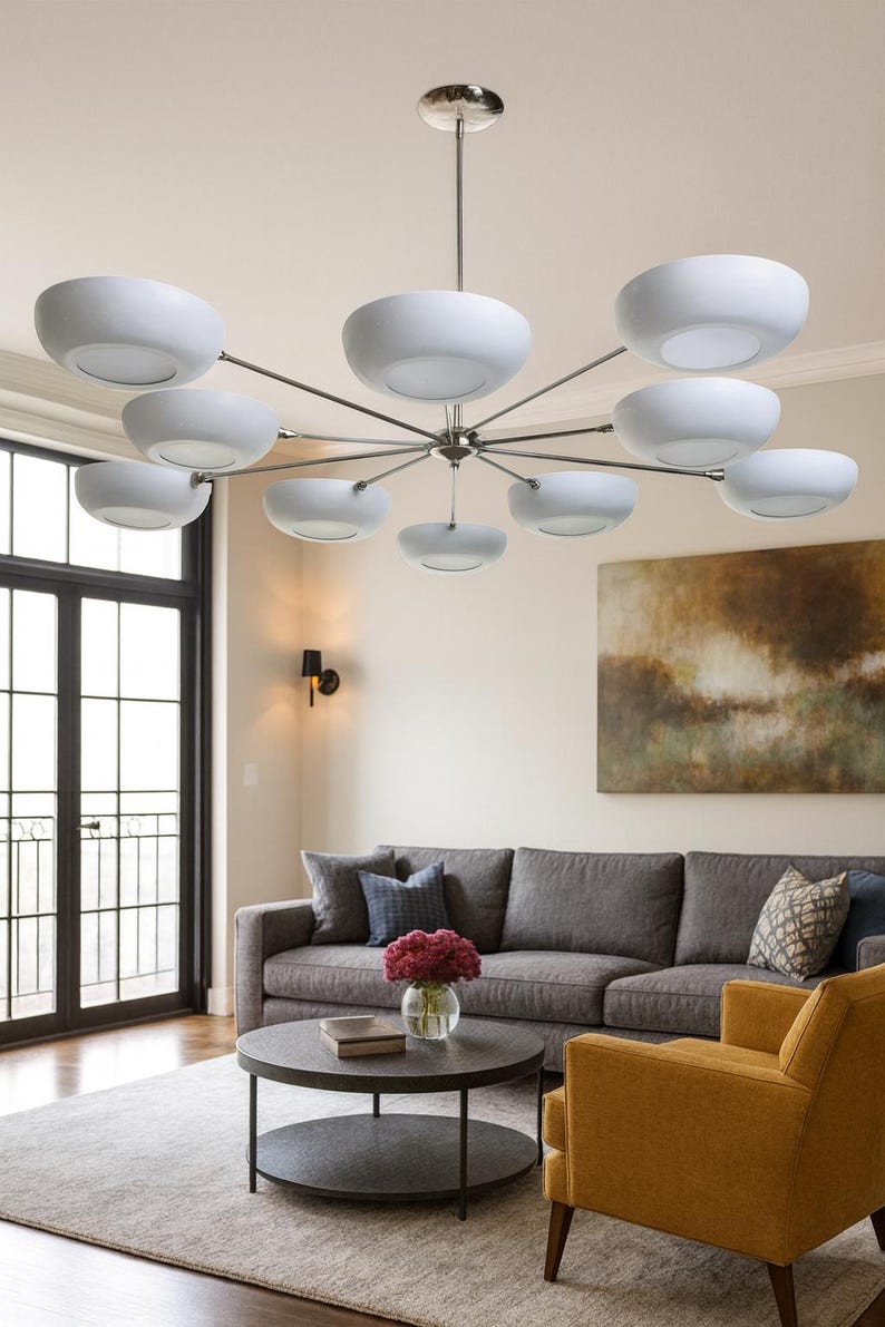 May include: A modern chandelier with multiple white bowl-shaped lights suspended from a central fixture. The lights are arranged in a circular pattern, with a polished metal finish. The chandelier hangs above a living room with a gray sofa and yellow armchair.