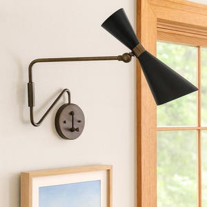 May include: A black and brass wall sconce with a conical shade. The adjustable arm and circular base are brass-toned. The sconce is mounted on a white wall, near a window with a framed landscape painting.