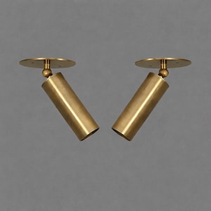 May include: Two brass-colored, cylindrical spotlights with a circular base and a ball joint. The spotlights are angled and appear to be made of metal. The background is a solid, neutral gray.