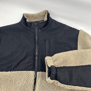 May include: A black and beige fleece jacket with a full zip. The jacket has a high collar and a contrasting texture between the smooth black fabric and the beige fleece material. A pocket is visible on the chest.