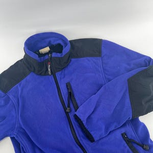 May include: A blue fleece jacket with black shoulder and collar accents. The jacket has a full-length zipper and two zippered pockets. The collar is a high, stand-up style. The jacket is made of a soft, warm material.