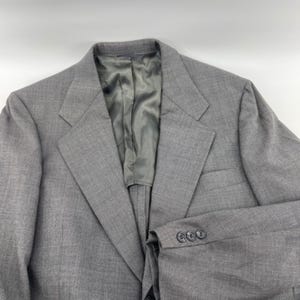 May include: A gray blazer with a notched lapel and three buttons on the sleeve. The interior lining is a light gray color. The blazer is made of a textured fabric.