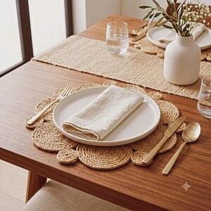 Peut inclure: Un set de table rond en jute tressé à bord festonné, surmonté d'une assiette en céramique blanche avec une serviette en lin beige pliée. Une fourchette, un couteau et une cuillère dorés sont disposés sur une table en bois avec un chemin de table.
