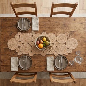 Peut inclure: Une table à manger rectangulaire en bois avec quatre chaises, dotée d'un chemin de table en jute tressé composé de disques circulaires. Un bol en céramique rempli de fruits frais assortis est placé au centre, entouré de deux couverts avec assiettes.