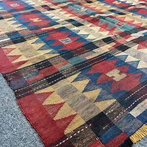 May include: A large, rectangular rug with a geometric pattern in red, blue, tan, and black. The rug features a repeating design of triangles and squares, with a fringed edge. The rug is made of woven fabric.