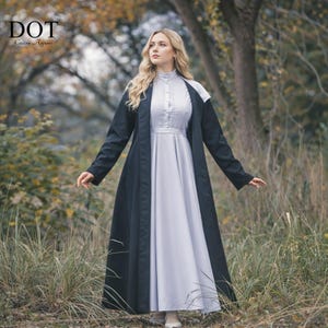 Pode incluir: Uma mulher vestindo um longo vestido cinza claro com gola alta e detalhes de botões, sob um longo casaco preto. O casaco tem detalhes brancos nos ombros. A imagem é ao ar livre com o texto "DOT Custom Apparels" no canto superior esquerdo.