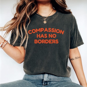 May include: Dark gray t-shirt with the orange text "COMPASSION HAS NO BORDERS." The shirt has a vintage, worn look. The model is wearing blue jeans and a gold necklace. The sleeves are short.