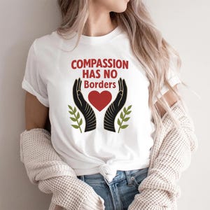 May include: White t-shirt with the text "COMPASSION HAS NO Borders" in red. The design includes black hands cradling a red heart, with green sprigs on either side. A simple, yet powerful message.