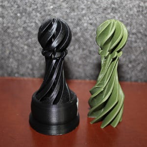 May include: Two decorative sculptures. One is black, the other is green. Both have a spiral design with a rounded top and a cylindrical base. The sculptures are displayed on a reddish-brown surface against a textured gray background.