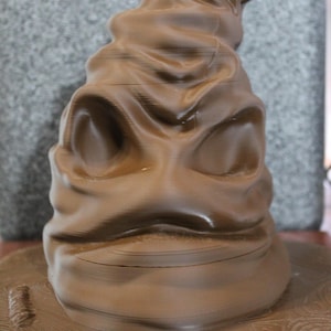 May include: A brown Sorting Hat, a wizarding hat from the Harry Potter series, with a wide brim and a wrinkled, expressive face. The hat is made of a dark brown material and has a textured appearance. The hat is a prop from the Harry Potter series.