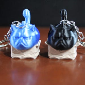 May include: Two keychains featuring a pug design, one blue and one black, each with a silver keyring. The pug heads are 3D-printed and sit atop a beige base. The background is dark.