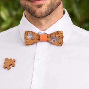 Op de afbeelding: Een houten vlinderdas met een puzzelstukjesontwerp en een oranje middenband. De vlinderdas is gemaakt van lichtbruin hout en wordt gecombineerd met een bijpassende reversspeld van een puzzelstukje. Gedragen met een wit overhemd.
