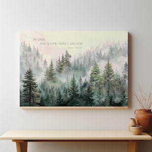 May include: A rectangular wall art print featuring a misty forest landscape with evergreen trees in shades of green and grey. The text "Be Still AND KNOW THAT I AM GOD Psalm 46:10" is printed in a dark font against the pale, foggy sky background.