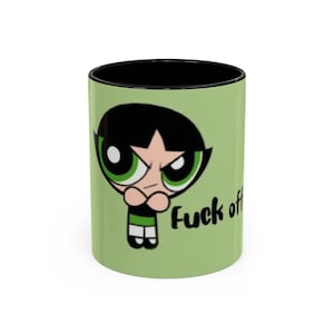 May include: A ceramic mug with a black interior and a light green exterior. The mug features a cartoon image of a girl with black hair and green eyes, with the words "Fuck off" printed in black.