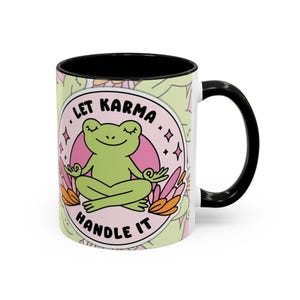 以下が含まれることがあります： 黒い内側とハンドルが付いた白いセラミックマグ。マグには、「Let Karma Handle It」という言葉が入ったピンク色の円の中に、ヨガのポーズをとる緑色のカエルが描かれています。背景には、薄緑色の花柄があります。