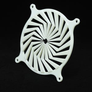 May include: A white, circular plastic vent cover with a geometric design. The cover has angled, chevron-shaped slots radiating from a central hole. Three small mounting tabs are evenly spaced around the perimeter.