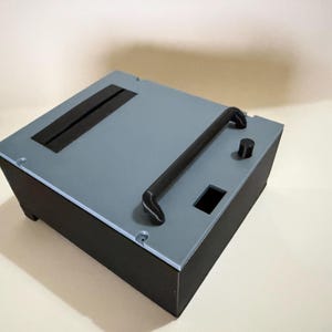 Può includere: Un contenitore elettronico rettangolare con base nera opaca e pannello superiore azzurro. Il pannello superiore presenta una lunga maniglia nera, una fessura rettangolare, un piccolo foro quadrato e una singola manopola di controllo nera.