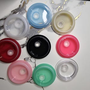 May include: An overhead shot displays nine colorful, circular lids. The lids are in various colors, including clear, blue, red, black, pink, green, and yellow. Each lid has a central, raised circular feature.