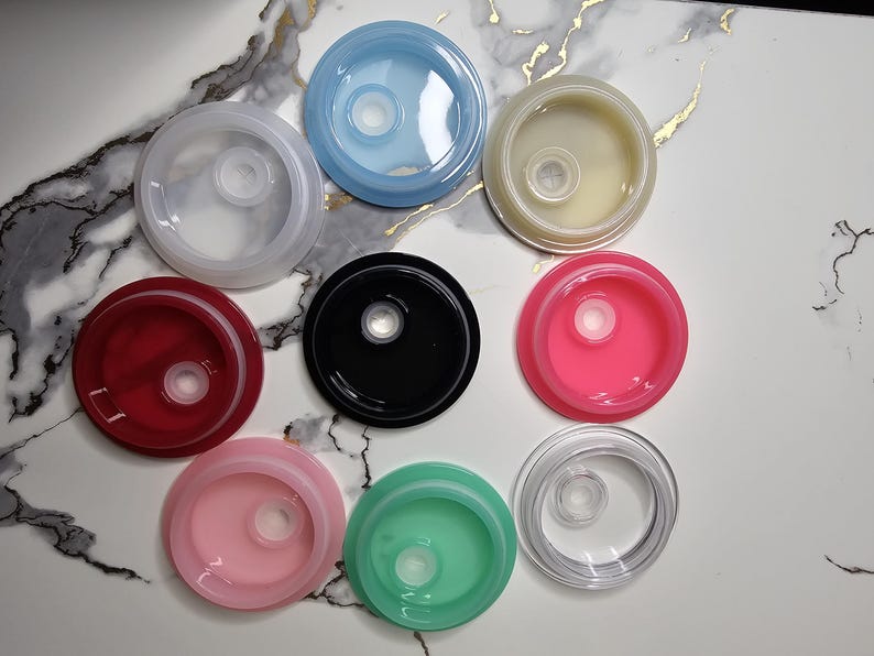 May include: An overhead shot displays nine colorful, circular lids. The lids are in various colors, including clear, blue, red, black, pink, green, and yellow. Each lid has a central, raised circular feature.