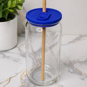 Pode incluir: Um copo de vidro transparente em forma de lata de refrigerante, com uma tampa de silicone azul vibrante com uma abertura central. Uma palhinha de madeira clara e reta está inserida através da tampa, estendendo-se verticalmente no copo.