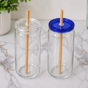 May include: Two clear glass drinking jars with lids and wooden straws. One jar has a clear lid, and the other has a blue lid. The jars are on a marble surface, with a white pot of greenery in the background.